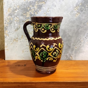 May include: A small, brown ceramic pitcher with a handle and a spout. The pitcher is decorated with yellow and green geometric patterns. The pitcher is sitting on a wooden surface.