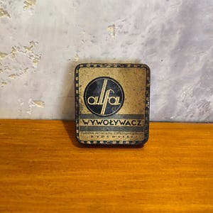 May include: A square, weathered metal tin with a gold and black finish. The lid features a circular logo with the text 'allfal', the word 'WYWOŁYWACZ', and the text 'FABRYKA ARTYKUŁÓW FOTOGRAFICZNYCH BYDGOSZCZ' printed in a clear, sans-serif font.