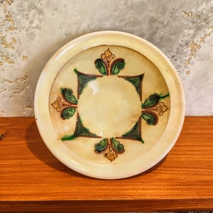 May include: A white ceramic plate with a green and brown floral design. The plate has a raised rim and a slightly concave centre.