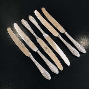 May include: Six silver butter knives with ornate handles. Each knife has a different inscription on the blade, including "G.S. 101", "G.S. 59", "G.S. 55", "G.S. 53", "G.S. 52", and "G.S. 51".