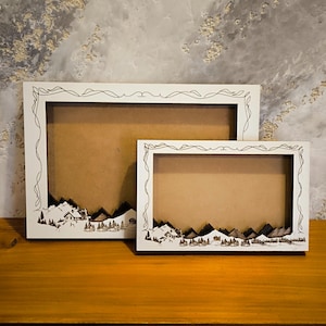 May include: Two white wooden picture frames with a decorative mountain scene design. The frames are nested inside each other, with the smaller frame in front of the larger frame.