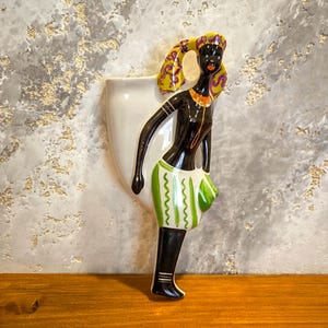 May include: A vintage ceramic wall pocket depicting a stylized African woman. The figure has black skin, a yellow head wrap with purple accents, and a green and white skirt. The wall pocket is white.