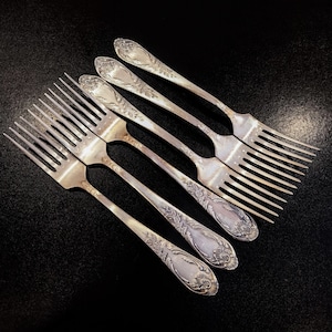 May include: Six antique silver forks arranged diagonally on a black surface. The forks feature ornate floral and scrollwork designs on the handles. The tines are long and slender, and the silver has a polished finish.