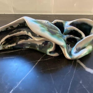 May include: Abstract ceramic sculpture with a flowing, organic form. The piece has a glossy finish with dark green, teal, and white colours. The sculpture's design is reminiscent of a folded or draped shape.