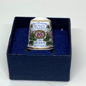 May include: A white porcelain thimble with a floral design and gold trim. The thimble features the text "Her Majesty Queen Elizabeth the Queen Mother" and the number "95".