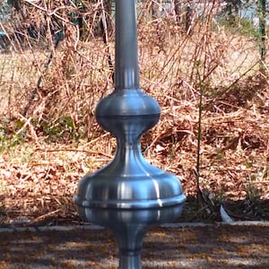 Puede incluir: Un remate de metal plateado con una punta puntiaguda y una base redondeada. El remate está diseñado para colocarse en la parte superior de un techo u otra estructura.