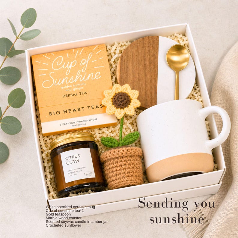 May include: A gift box containing a box of Cup of Sunshine herbal tea, a white speckled ceramic mug with a tan base, a gold teaspoon, a round marble and wood coaster, a scented soy candle in an amber jar, and a small crocheted sunflower in a pot.
