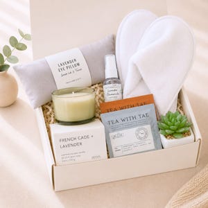 May include: A gift box containing a light gray lavender eye pillow, a pair of white slippers, a small glass candle, a spray bottle labeled Lavender Chamomile, a box of French Cade and Lavender soy wax candle, a tea packet labeled Tea With Tae, and a succulent.