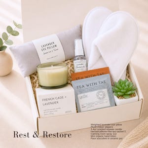 May include: A gift box containing a gray lavender eye pillow, white plush slippers, a 3.4oz French Cade and Lavender soy candle, a lavender chamomile room spray, two Tea With Tae herbal tea sachets, and a small faux succulent in a ceramic pot.