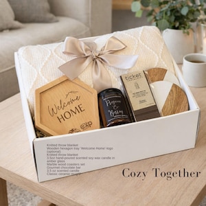 Puede incluir: Una caja de regalo que contiene una manta de punto color crema atada con una cinta beige, una bandeja de madera hexagonal con el texto Welcome HOME, una vela de vidrio &aacute;mbar de 3.5 oz etiquetada Prosecco & Berries, chocolate Ticket y posavasos.
