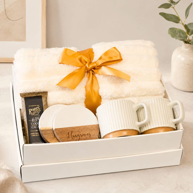 May include: A gift set in a white box containing a folded cream-colored plush blanket tied with a gold ribbon, two white ribbed ceramic mugs with tan bases, two round white and wood coasters with the name Hargroves, and a bar of dark chocolate.