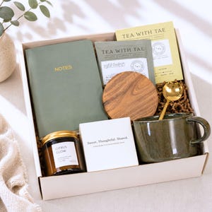 Puede incluir: Una caja de regalo que contiene un cuaderno verde salvia con la palabra NOTES, una bolsita de t&eacute; Tea With Tae, un posavasos redondo de madera, una cuchara dorada, una taza de cer&aacute;mica verde oscuro, una vela Citrus Glow y una caja blanca con el texto Sweet. Thoughtful. Shared.