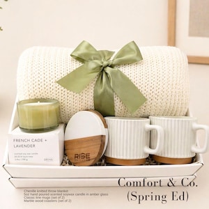 Puede incluir: Un set de regalo que incluye una manta de chenilla color crema atada con una cinta verde salvia, una vela de cera de soja de 100g, dos tazas de cer&aacute;mica blanca acanalada y un posavasos redondo de m&aacute;rmol y madera.