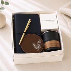 May include: A gift box containing a navy blue journal, gold pen, brown coaster with a white leaf design, a black travel mug with a wood-grain band, and a white box with text. The box is white with a cream interior.