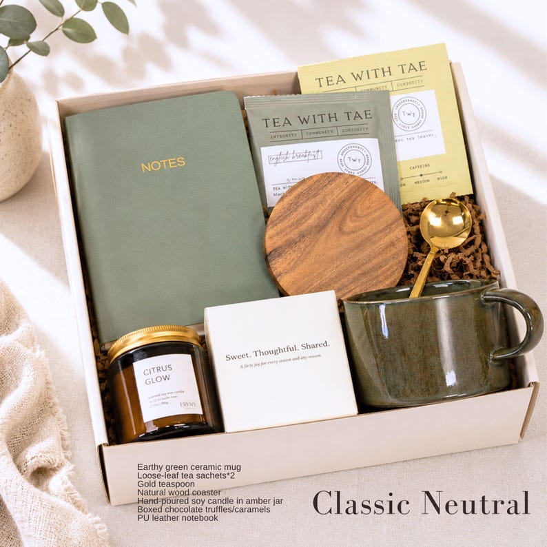 K&ouml;nnte beinhalten: Eine Geschenkbox mit einer gr&uuml;nen Keramiktasse, Teesachets, einem goldenen Teel&ouml;ffel, einem Holzuntersetzer, einer Kerze, einem Notizbuch und Pralinen. Die Box tr&auml;gt die Aufschrift "Classic Neutral" und enth&auml;lt den Text "Sweet. Thoughtful. Shared."