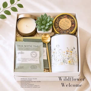May include: A gift box containing a 3.5 oz scented soy wax candle in a gold tin, a faux succulent in a white ceramic pot, a tin of botanical facial steam, three tea sachets labeled Tea With Tae, a gold teaspoon, and a white insulated tumbler.