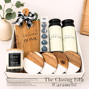 Puede incluir: Una caja de regalo con una tabla de madera con el texto 'Welcome HOME' y un lazo a cuadros blanco y negro, chocolates JACEK, dos vasos aislados color crema de 11 oz con 'HOMES BY NEW ERA', cuatro posavasos de m&aacute;rmol y madera, y una vela de 5,5 oz.