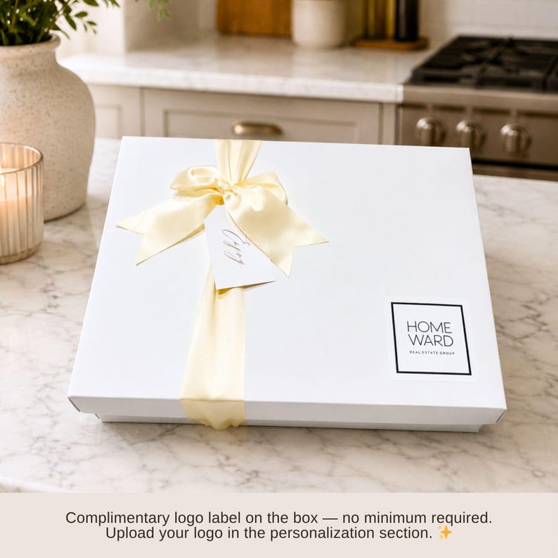 Puede incluir: Una caja de regalo cuadrada de color blanco con un lazo de sat&eacute;n amarillo p&aacute;lido en la parte superior. Una peque&ntilde;a etiqueta rectangular blanca est&aacute; sujeta al lazo. Una pegatina cuadrada con borde negro muestra el texto HOME WARD REAL ESTATE GROUP.