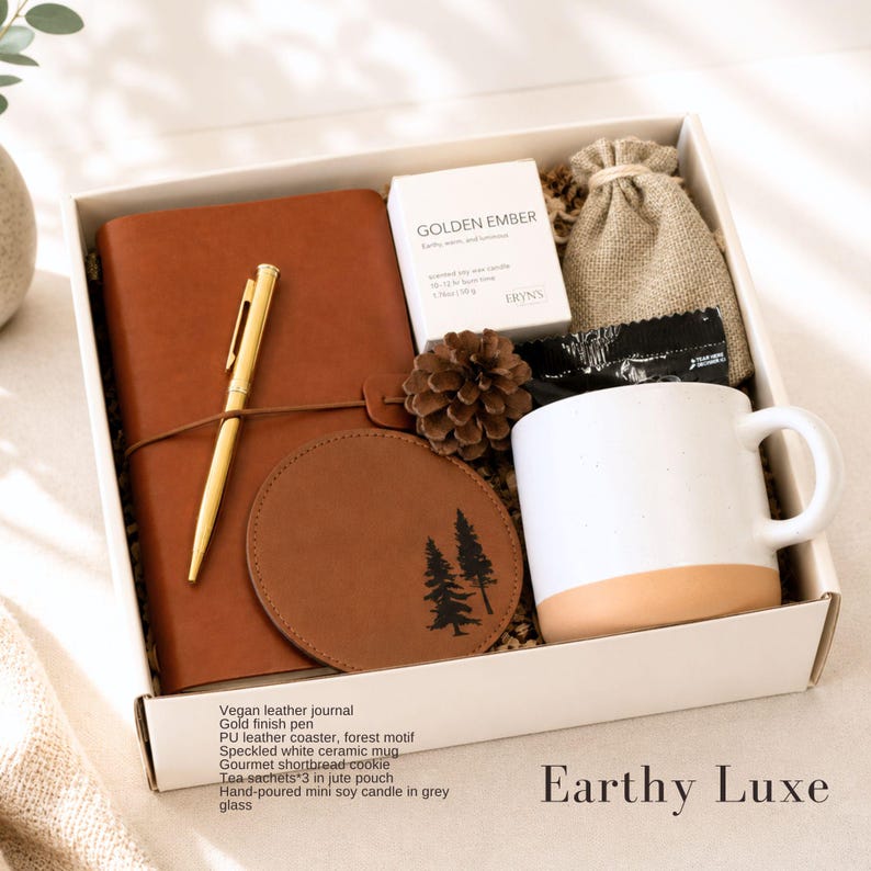 May include: A gift box containing a brown vegan leather journal, a gold finish pen, a round brown PU leather coaster with a forest motif, a speckled white ceramic mug, a small jute pouch, a pinecone, and a box labeled Golden Ember.
