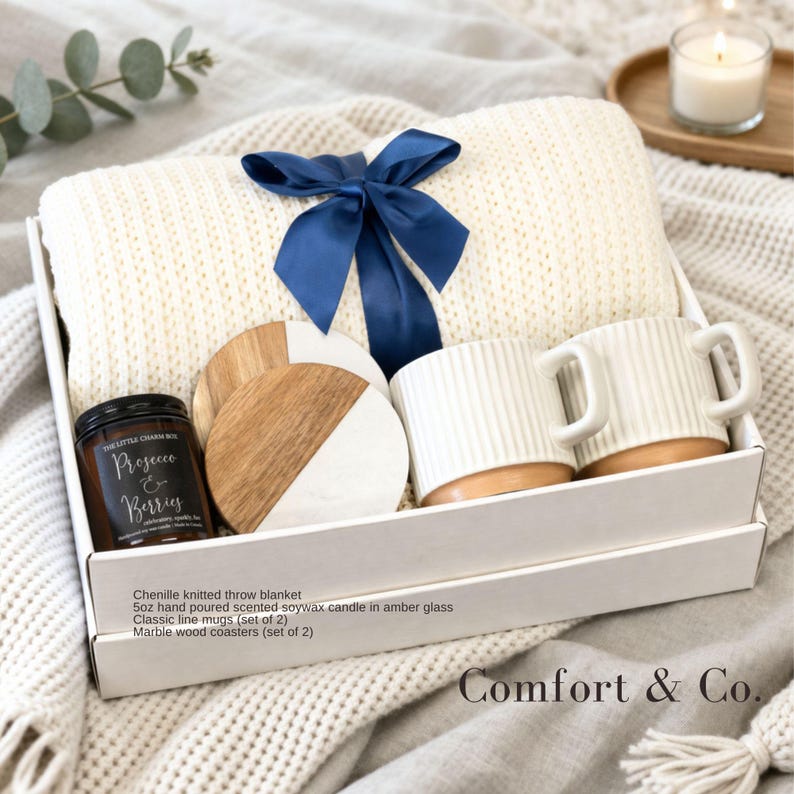 May include: A gift set in a white box containing a cream chenille knitted throw blanket tied with a navy blue ribbon, two white ribbed ceramic mugs, two round marble and wood coasters, and a 5oz Prosecco & Berries scented soy wax candle.