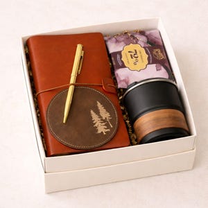 Può includere: Una confezione regalo contenente un diario in pelle marrone, una penna dorata, un sottobicchiere marrone con un disegno di albero, una tazza da viaggio nera con una fascia effetto legno e una barretta di cioccolato fondente. Gli articoli sono in una scatola bianca.