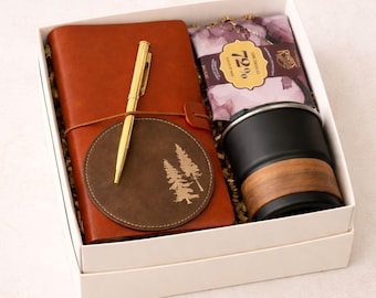 Puede incluir: Una caja de regalo que contiene un diario de cuero marrón, un bolígrafo dorado, un posavasos marrón con un diseño de árbol, una taza de viaje negra con una banda de vetas de madera y una barra de chocolate negro. Los artículos están en una caja blanca.