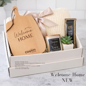 Puede incluir: Una caja de regalo que contiene una manta de punto de color claro atada con una cinta, una tabla de madera grabada con Welcome HOME y Clayton Homes, una vela de cera de soja de 142g, una suculenta artificial en maceta blanca y chocolate.