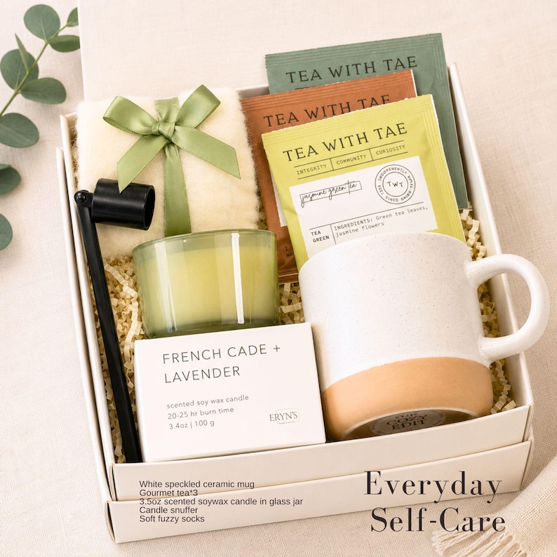 K&ouml;nnte beinhalten: Eine wei&szlig;e Geschenkbox mit Artikeln zur Selbstpflege. Sie enth&auml;lt eine gesprenkelte Keramiktasse, eine Duftkerze aus Sojawachs, Teebeutel, einen Kerzenl&ouml;scher und weiche, flauschige Socken. Die Box ist mit einem gr&uuml;nen Band gebunden und tr&auml;gt den Text "Everyday Self-Care".
