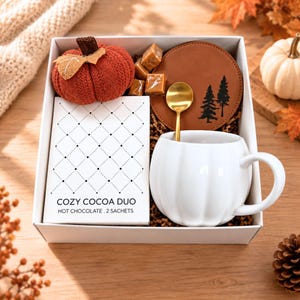 Può includere: Una confezione regalo contenente una zucca arancione lavorata a maglia, una scatola bianca con etichetta COZY COCOA DUO HOT CHOCOLATE 2 SACHETS, cubetti di caramello, un sottobicchiere rotondo in pelle marrone con pini neri, un cucchiaio oro e una tazza.