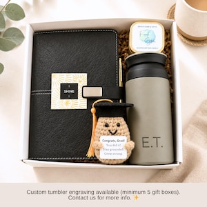 K&ouml;nnte beinhalten: Eine Geschenkbox mit einem schwarzen Notizbuch, einem grauen Becher mit der Gravur "E.T.", einer Kerze und einer H&auml;kel-Abschluss-Puppe. Das Notizbuch hat ein "SHINE"-Etikett. Die Puppe hat eine Abschlusskappe und ein Etikett mit "Congrats, Grad! You did it! Stay grounded. Grow strong."