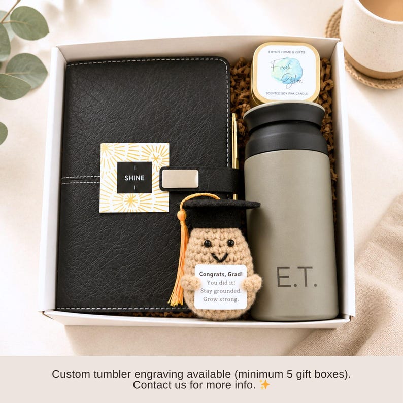 K&ouml;nnte beinhalten: Eine Geschenkbox mit einem schwarzen Notizbuch, einem grauen Becher mit der Gravur "E.T.", einer Kerze und einer H&auml;kel-Abschluss-Puppe. Das Notizbuch hat ein "SHINE"-Etikett. Die Puppe hat eine Abschlusskappe und ein Etikett mit "Congrats, Grad! You did it! Stay grounded. Grow strong."