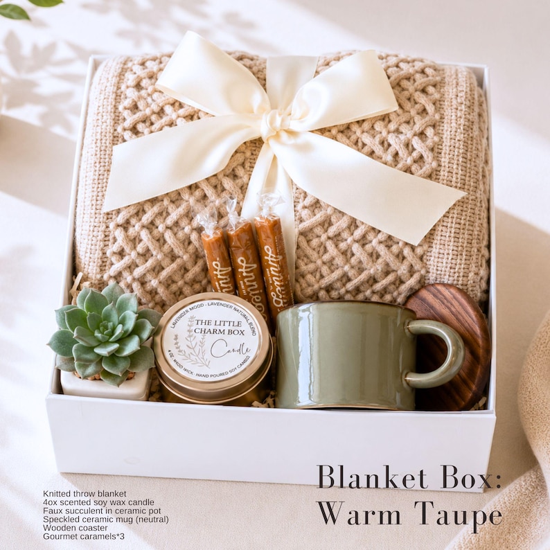 May include: A white gift box containing a beige knitted throw blanket tied with a cream ribbon, three gourmet caramels, a small faux succulent in a white ceramic pot, a round candle tin labeled The Little Charm Box, and a green ceramic mug.