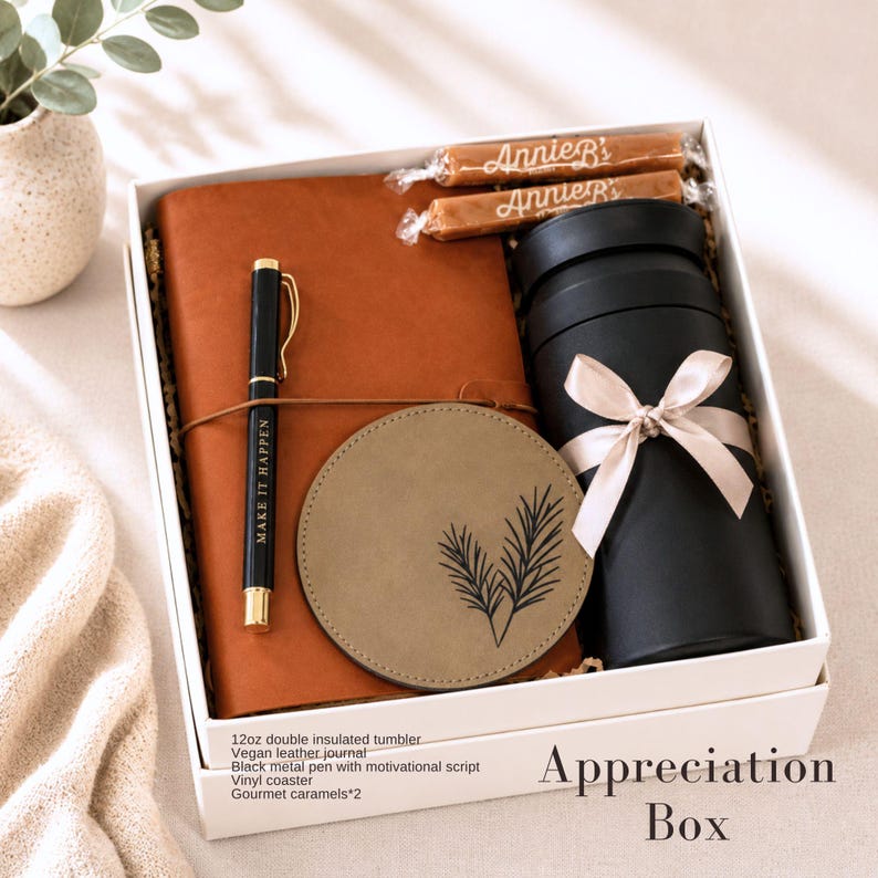 May include: A gift box containing a tan vegan leather journal, a black metal pen with the text MAKE IT HAPPEN, a round tan coaster with a leaf design, a black 12oz double insulated tumbler with a beige ribbon, and two Annie B's gourmet caramels.