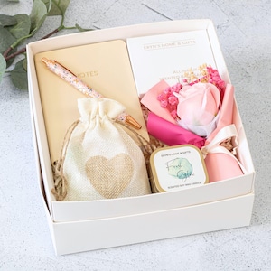 May include: A white gift box containing a beige notebook, a pen, a drawstring bag, a candle, and a floral arrangement. The notebook and pen have a pink and gold design. The bag features a heart. The candle is in a gold tin.