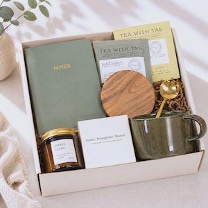 May include: A gift box containing a sage green notebook with the word NOTES, a Tea With Tae tea packet, a round wooden coaster, a gold spoon, a dark green ceramic mug, a Citrus Glow candle in a glass jar, and a white box labelled Sweet. Thoughtful. Shared.