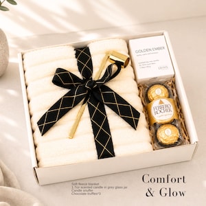 K&ouml;nnte beinhalten: Geschenkbox mit einer weichen Fleecedecke, einer Duftkerze, einem Kerzenl&ouml;scher und Schokoladentr&uuml;ffeln. Die Box ist mit einem schwarzen Band mit goldenen Diamantakzenten gebunden. Der Text "Comfort & Glow" ist sichtbar.