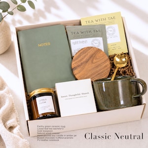 May include: A gift box containing a sage green notebook with the word NOTES, two tea packets labeled TEA WITH TAE, a round wooden coaster, a gold teaspoon, a dark green ceramic mug, a candle in an amber jar labeled CITRUS GLOW, and a white box.