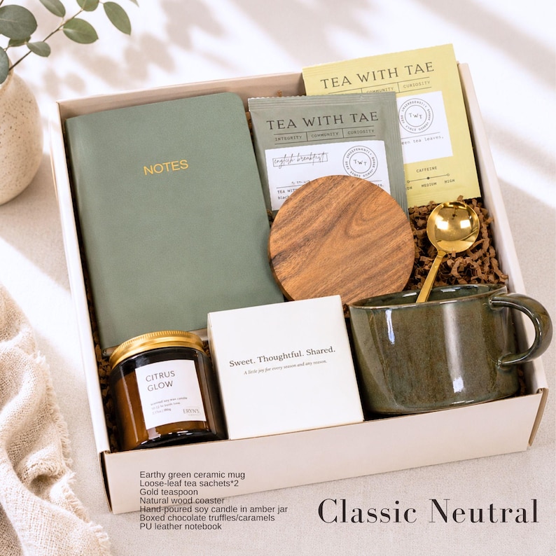 May include: A gift box containing a sage green notebook with the word NOTES, two tea packets labeled TEA WITH TAE, a round wooden coaster, a gold teaspoon, a dark green ceramic mug, a candle in an amber jar labeled CITRUS GLOW, and a white box.
