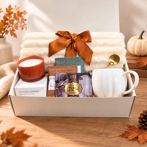 Può includere: Una confezione regalo contenente una coperta testurizzata color crema legata con un nastro arancione bruciato, una tazza bianca a forma di zucca, un cucchiaio dorato, una candela arancione bruciato etichettata Golden Ember, una bustina di tè e una tavoletta di cioccolato fondente al 72%.