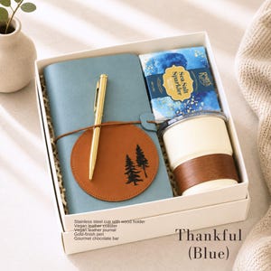 K&ouml;nnte beinhalten: Ein Geschenkset in einer wei&szlig;en Box enth&auml;lt ein blaues Notizbuch, einen goldenen Stift, einen braunen Untersetzer mit Baumdesign, einen wei&szlig;en Edelstahlbecher mit Holzhalter und einen Gourmet-Schokoriegel. Der Text "Thankful (Blue)" ist sichtbar.