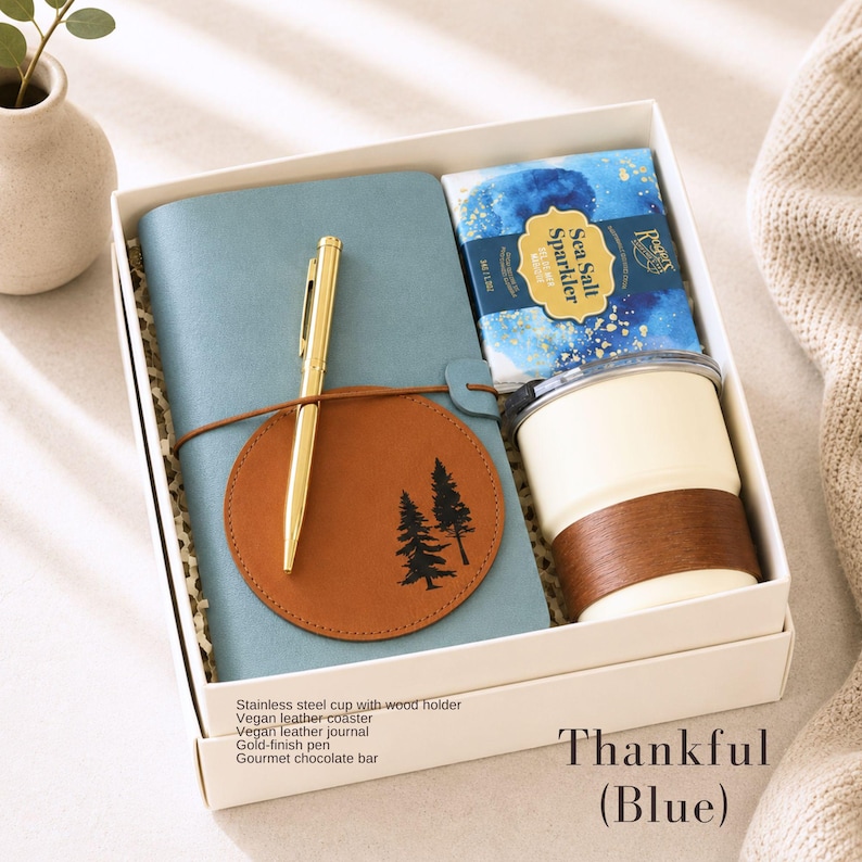K&ouml;nnte beinhalten: Ein Geschenkset in einer wei&szlig;en Box enth&auml;lt ein blaues Notizbuch, einen goldenen Stift, einen braunen Untersetzer mit Baumdesign, einen wei&szlig;en Edelstahlbecher mit Holzhalter und einen Gourmet-Schokoriegel. Der Text "Thankful (Blue)" ist sichtbar.