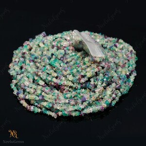 May include: A strand of multi-colored chip gemstone beads. The beads are a mix of green, blue, purple, and white. The beads are small and irregular in shape.