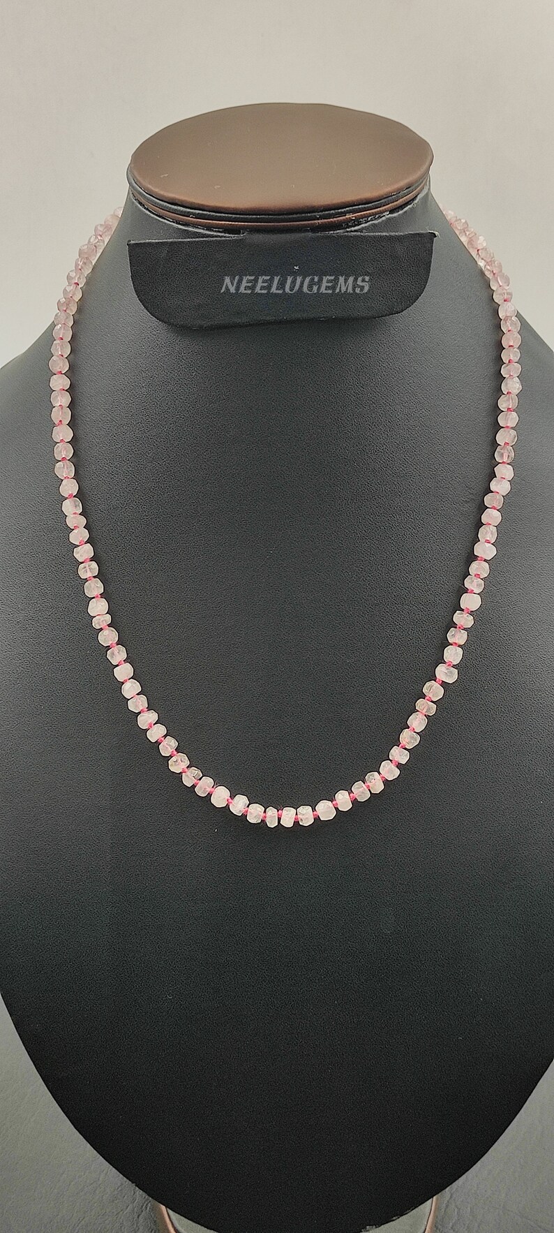 May include: A pink rose quartz beaded necklace with a pink thread. The necklace is displayed on a black mannequin bust. The text "NEELUGEMS" is visible on the bust.