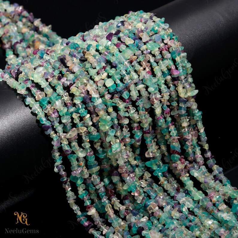 May include: A strand of multi-colored chip beads. The beads are a mix of green, blue, purple, and white. The beads are small and irregular in shape. The beads are strung on a clear thread.