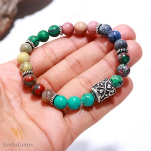 Puede incluir: Un brazalete de cuentas multicolores con un dije de plata. Las cuentas son verdes, amarillas, rojas, rosas, azules y turquesas. El dije tiene un diseño floral.