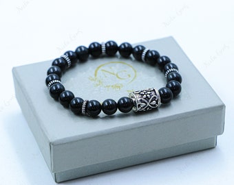 May include: A black beaded bracelet with a silver charm featuring an ornate design. The bracelet is displayed on a light gray box.