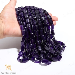 Könnte beinhalten: Eine Kette aus polierten, rechteckigen Amethystperlen. Die Perlen sind tiefviolett und wirken glatt und glänzend. Die Perlen werden in einer Hand vor weißem Hintergrund gehalten.