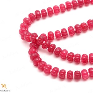 May include: Close-up of a strand of red gemstone beads. The beads are a deep, rich red color and have a carved, textured surface. The beads are strung together on a white string. The image is well-lit and the beads are in sharp focus.