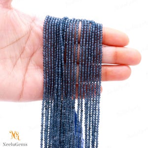 Peut inclure: Un ensemble de petites perles bleues foncées à facettes. Les perles sont tenues dans une main sur un fond blanc. Les perles sont enfilées ensemble, formant plusieurs brins. Le logo "NeeluGems" est dans le coin inférieur gauche.
