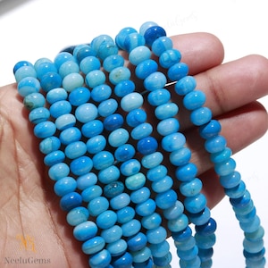 May include: Strands of blue gemstone beads in varying shades of light and dark blue. The beads are oval-shaped and strung together, creating a textured and colorful display. The beads are held in a hand.